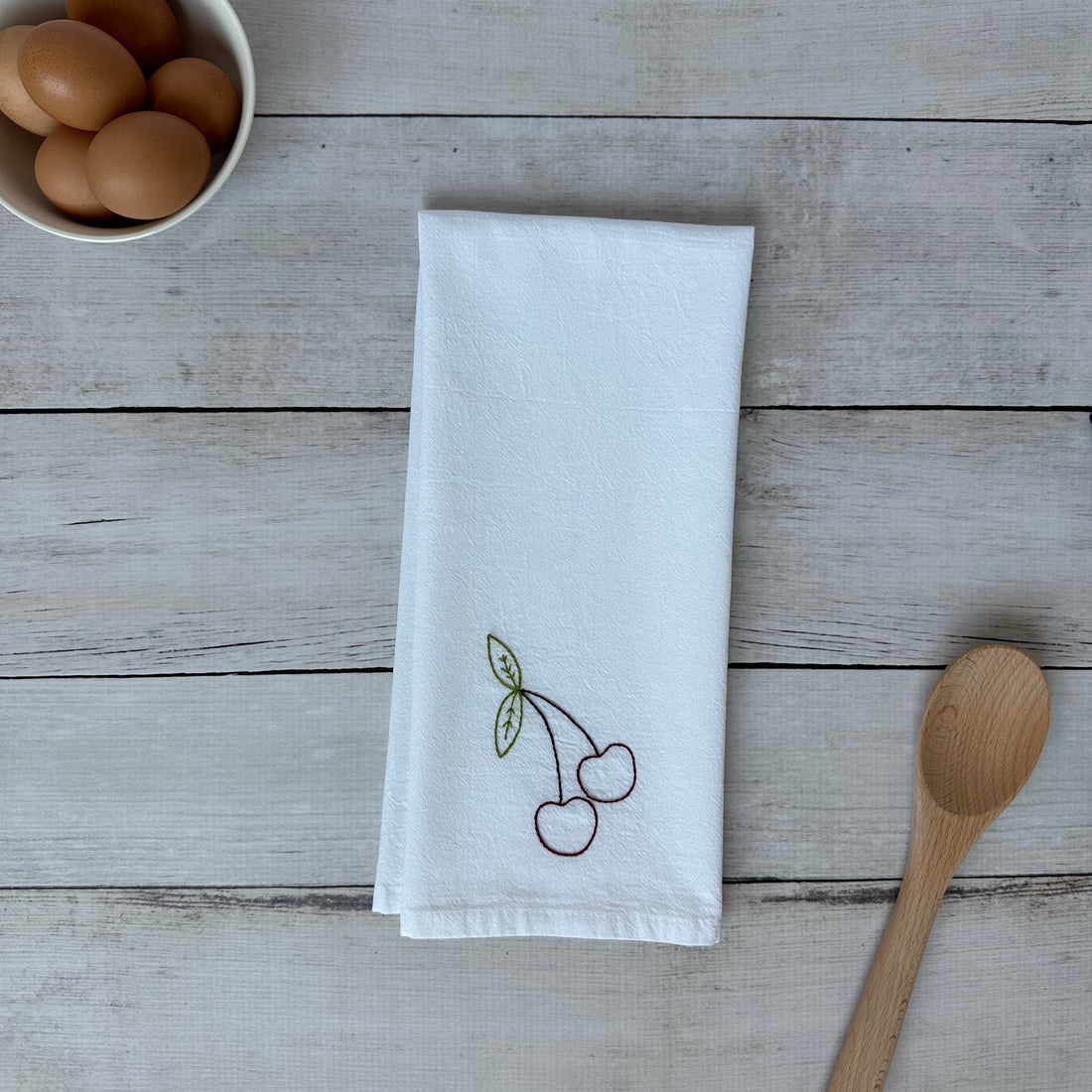 White tea towel with embroidered cherry design on a wooden surface with eggs and a wooden spoon.