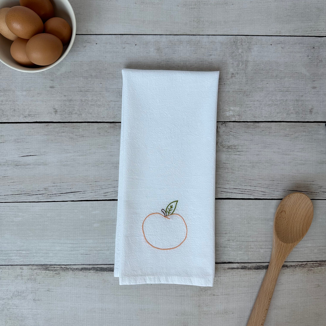 White tea towel with embroidered peach  design on a wooden surface with eggs and a spoon.