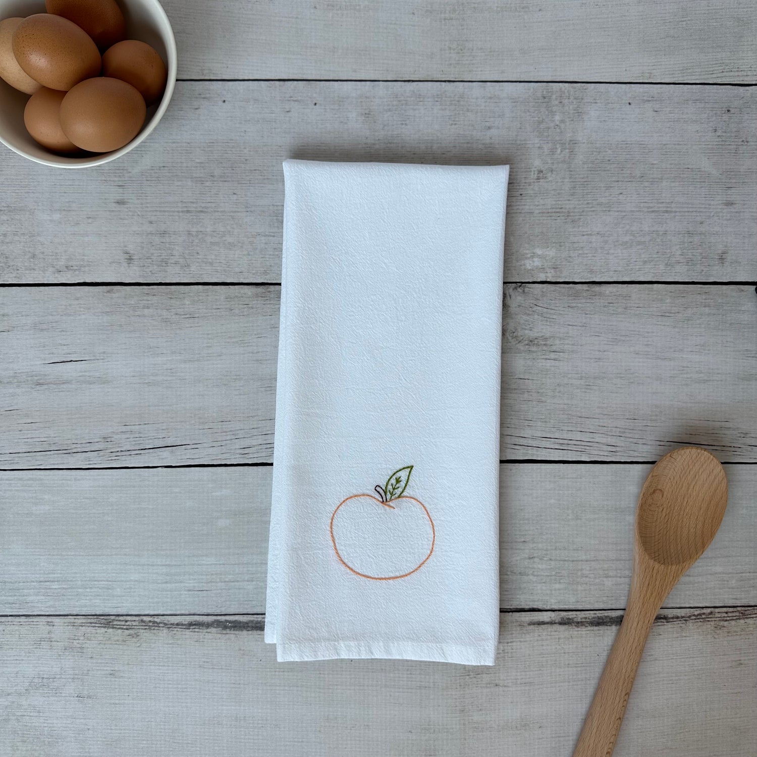 White tea towel with embroidered peach  design on a wooden surface with eggs and a spoon.