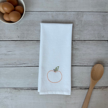 White tea towel with embroidered peach  design on a wooden surface with eggs and a spoon.