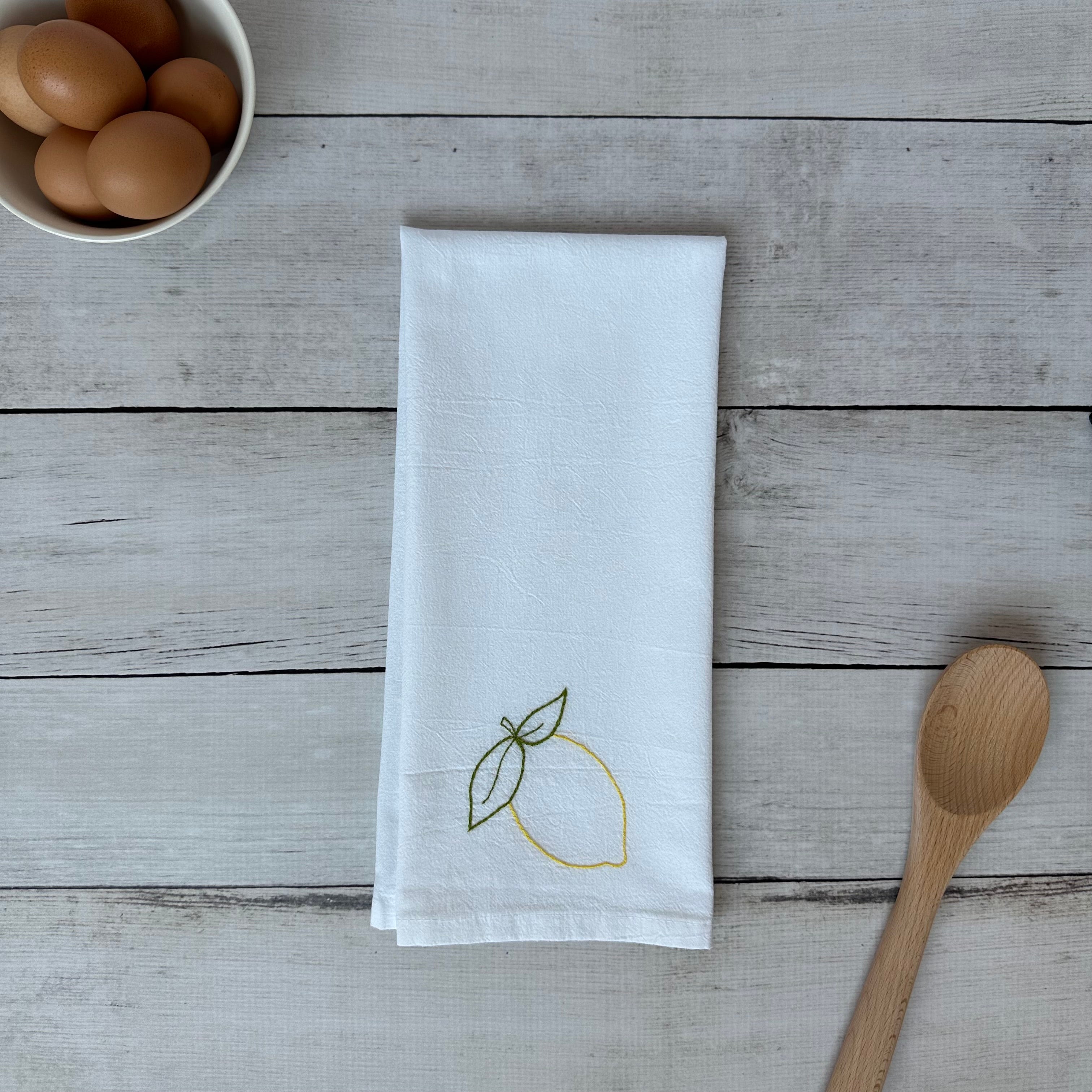 White tea towel with lemon embroidery on a wooden surface with eggs and a spoon.