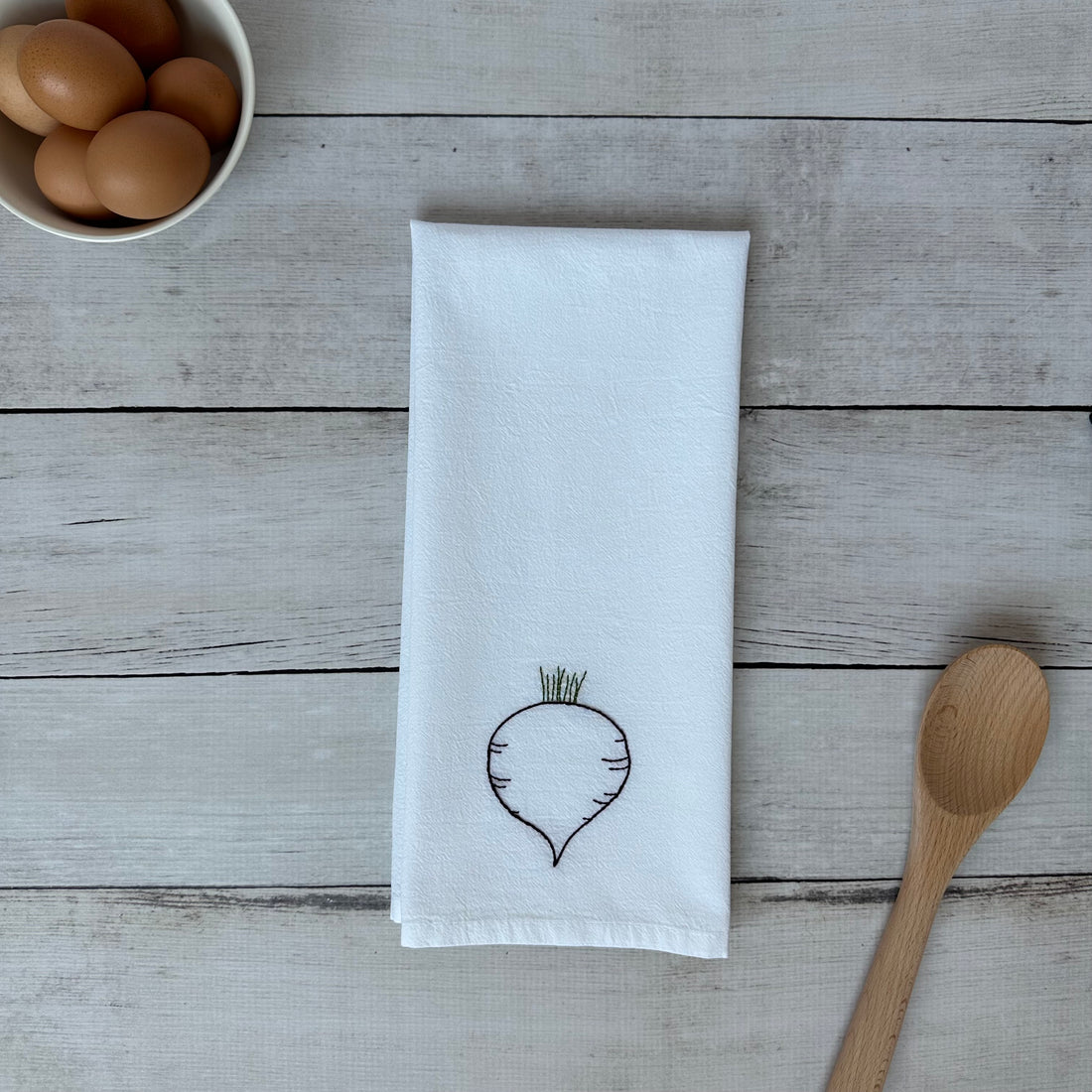 White tea towel with embroidered beet design on a wooden surface with eggs and a spoon.