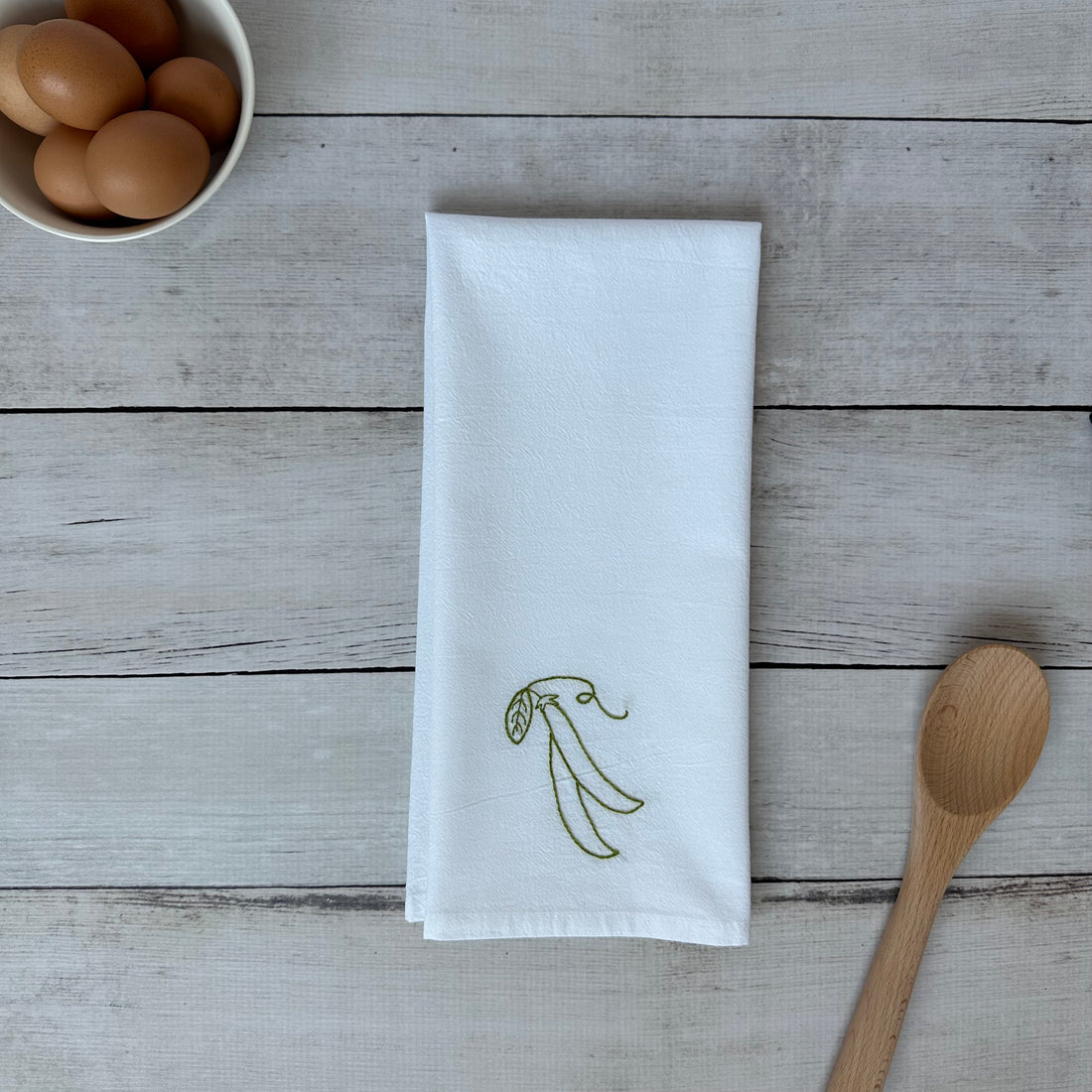 White tea towel with embroidered snap peas design on a wooden surface with eggs and a spoon.