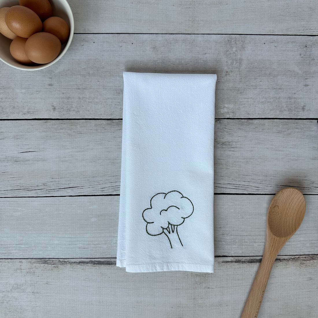 White tea towel with embroidered broccoli design on a wooden surface with eggs and a wooden spoon.