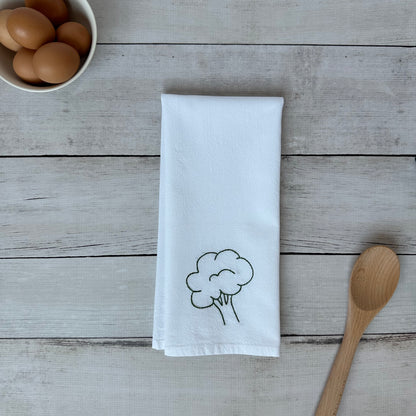 White tea towel with embroidered broccoli design on a wooden surface with eggs and a wooden spoon.