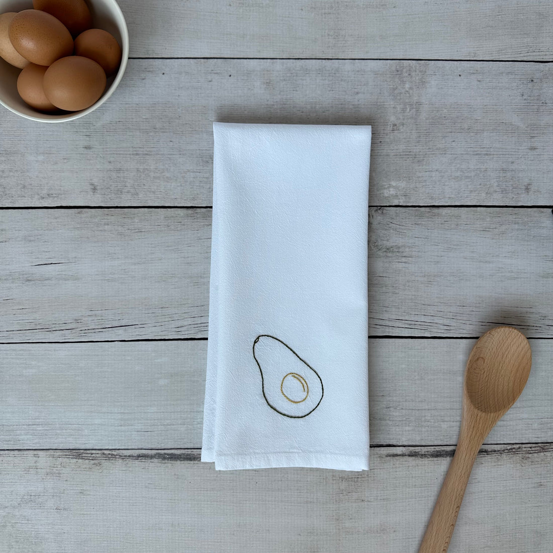 White tea towel with embroidered avocado design on a wooden surface with eggs and a spoon.