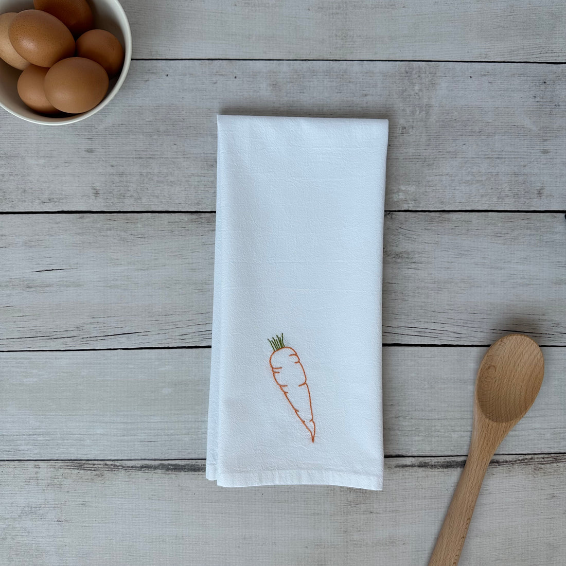 White tea towel with embroidered carrot design on a wooden surface with eggs and a spoon.