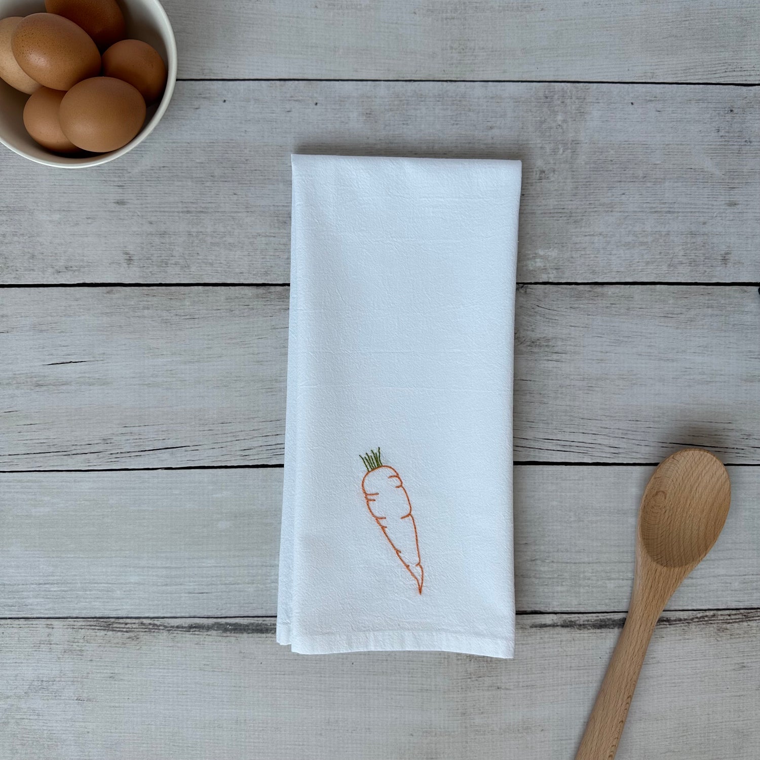 White tea towel with embroidered carrot design on a wooden surface with eggs and a spoon.