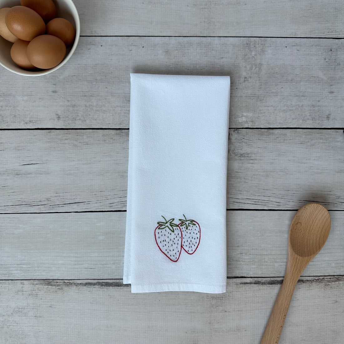 White tea towel with embroidered strawberry design on a wooden surface with eggs and a wooden spoon.