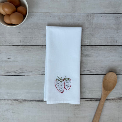 White tea towel with embroidered strawberry design on a wooden surface with eggs and a wooden spoon.