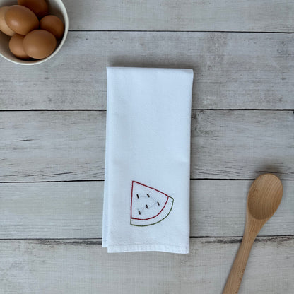 White tea towel with embroidered watermelon design on a wooden surface with eggs and a spoon.