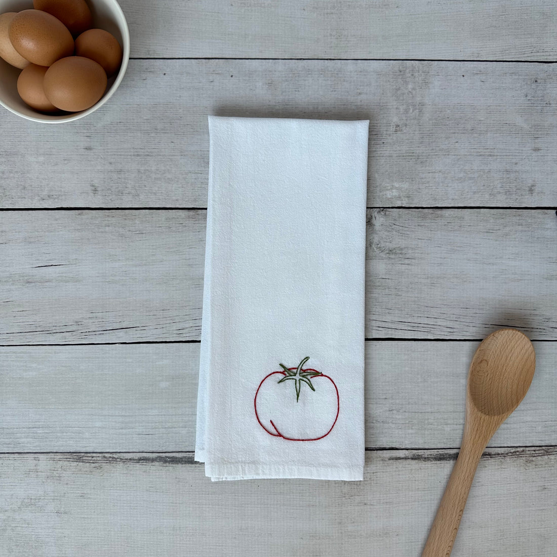 White tea towel with tomato embroidery on a wooden surface with eggs and a spoon.