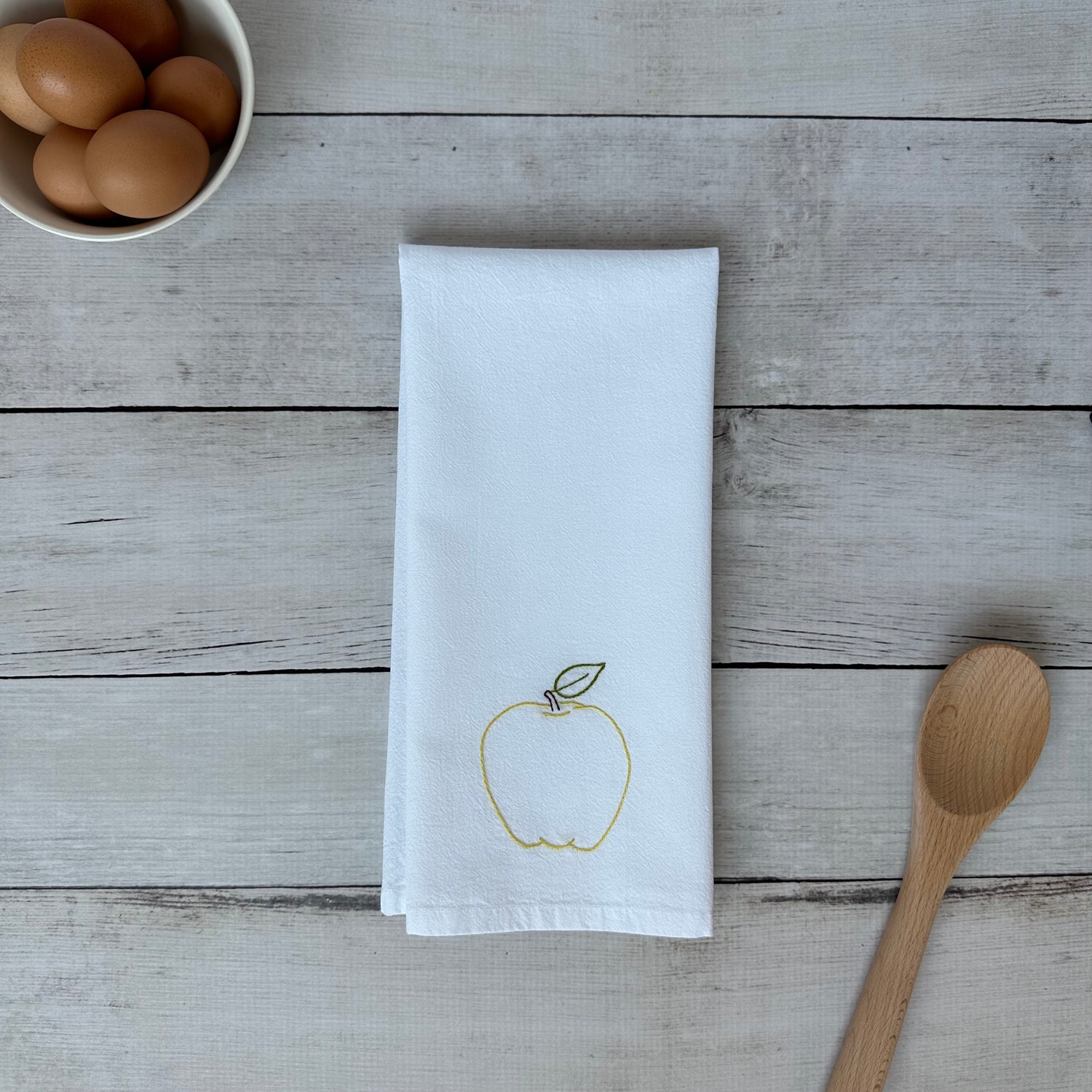 White tea towel with an embroidered yellow apple design on a wooden surface with eggs and a spoon
