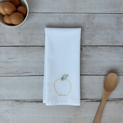 White tea towel with an embroidered yellow apple design on a wooden surface with eggs and a spoon