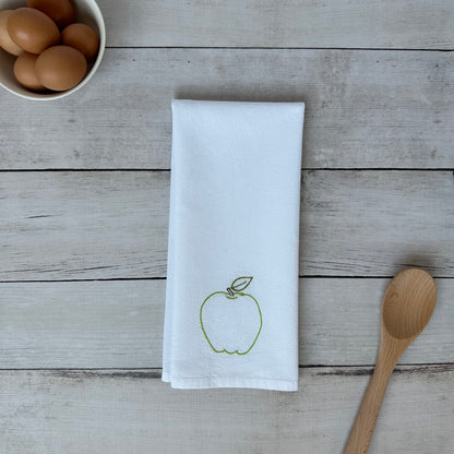 White tea towel with embroidered green apple design on a wooden surface with eggs and a wooden spoon.