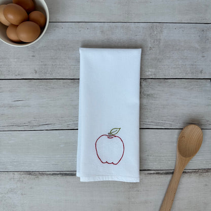White tea towel with an embroidered red apple design on a wooden surface with eggs and a wooden spoon.