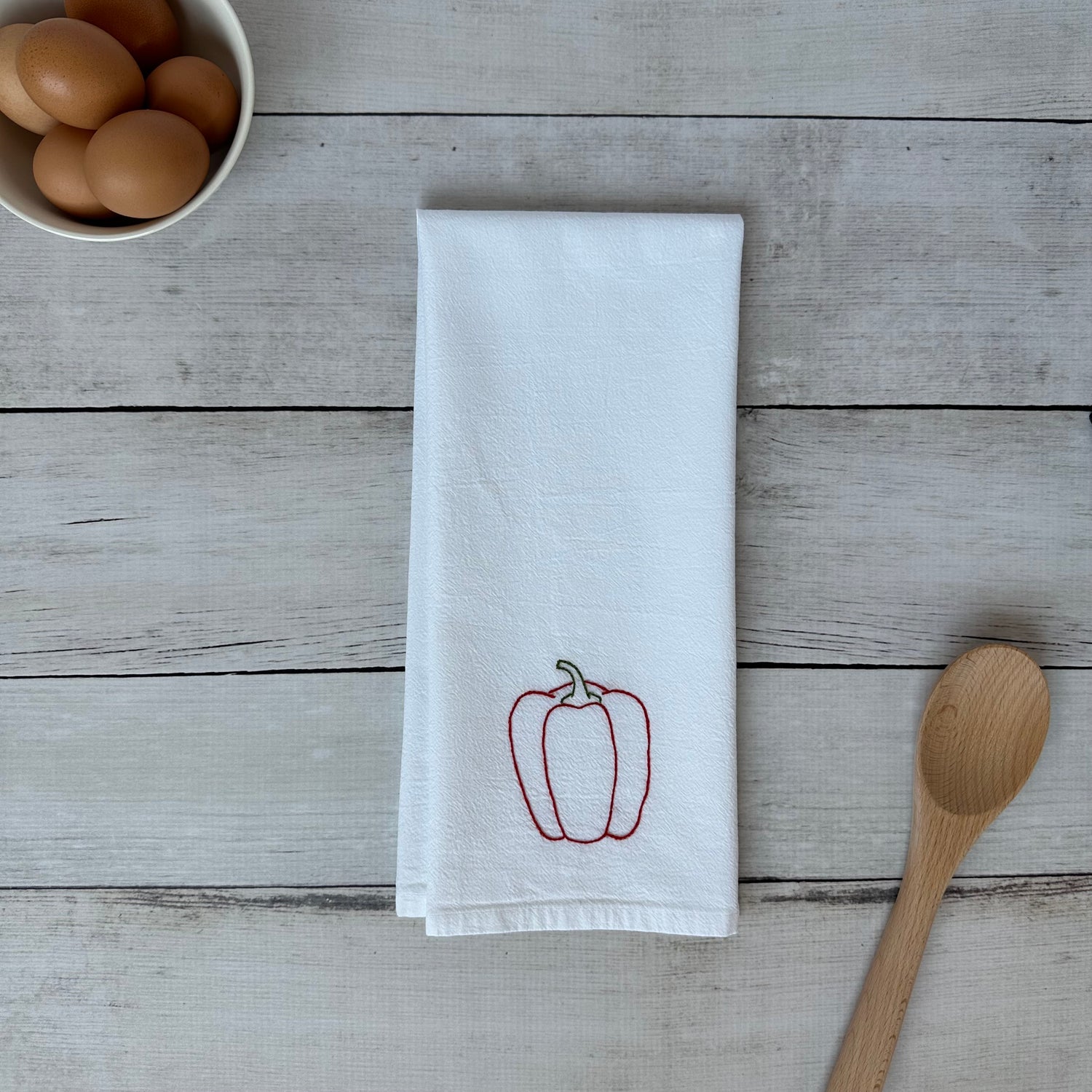 White tea towel with embroidered red bell pepper design on a wooden surface with eggs and a spoon.