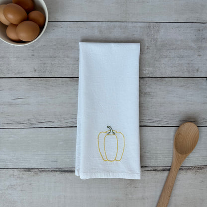 White tea towel with embroidered yellow bell pepper design on a wooden surface with eggs and a spoon.