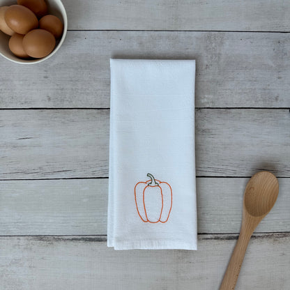 White tea towel with embroidered orange bell pepper design on a wooden surface with eggs and a spoon.