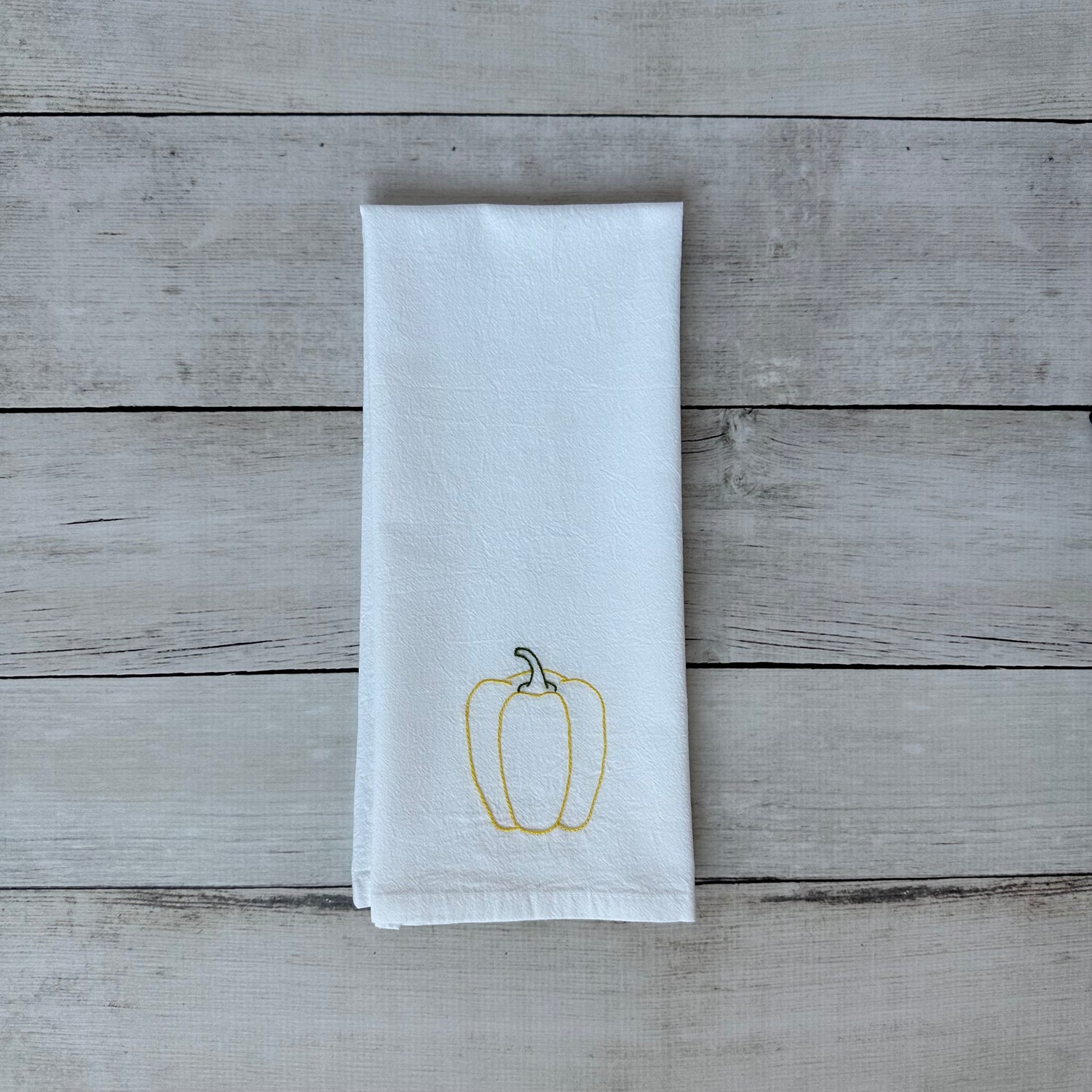 White tea towel with embroidered yellow bell pepper design on a wooden surface
