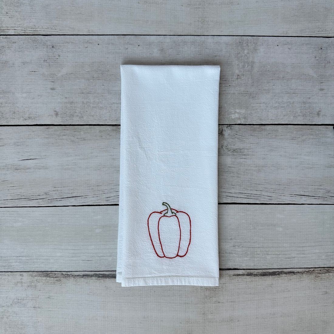 White tea towel with embroidered red bell pepper design on a wooden surface