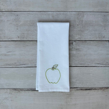 White tea towel with embroidered green apple design on a wooden surface