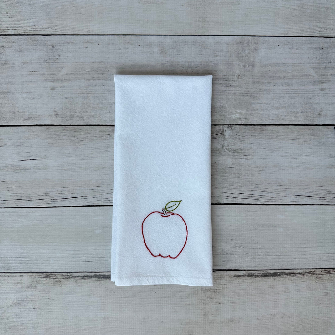 White tea towel with an embroidered red apple design on a wooden surface