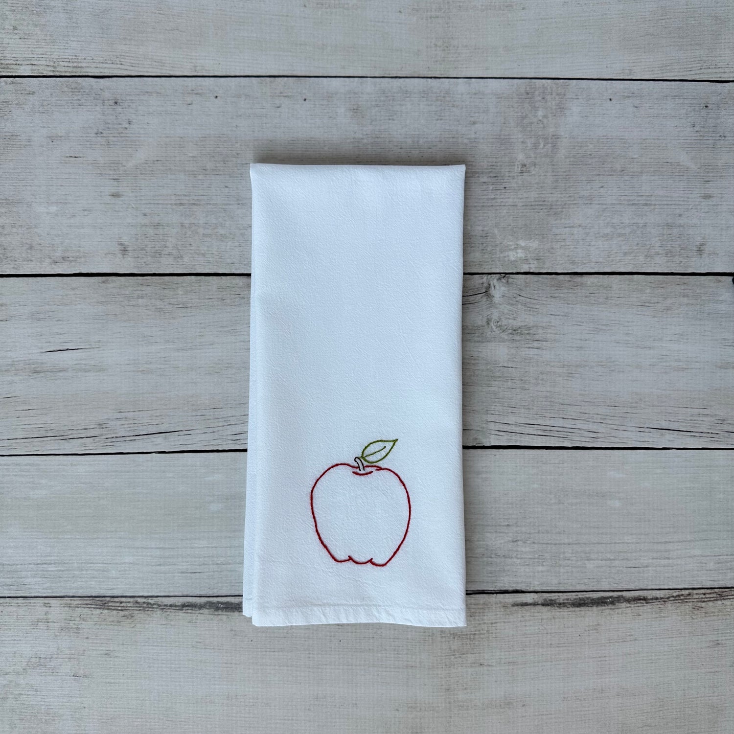 White tea towel with an embroidered red apple design on a wooden surface