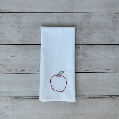 White tea towel with an embroidered red apple design on a wooden surface