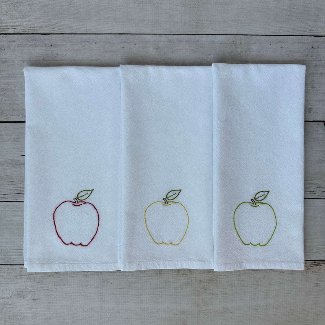 Three white tea towels with embroidered apple designs on a wooden surface