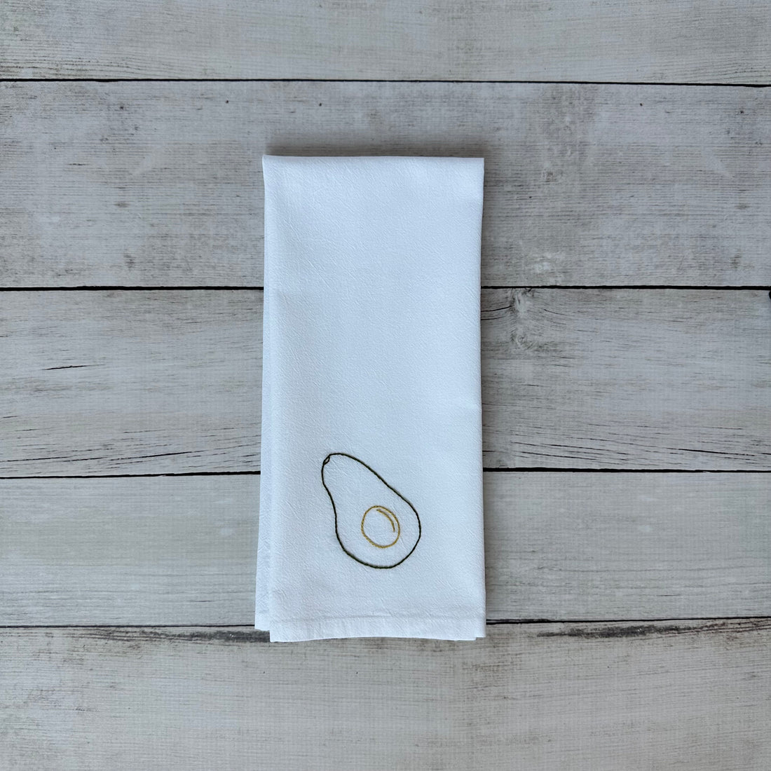 White tea towel with embroidered avocado design on a wooden surface