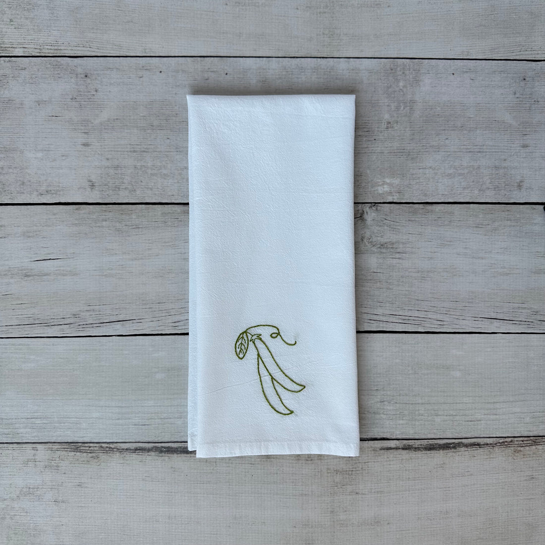 White tea towel with embroidered snap peas on a wooden surface