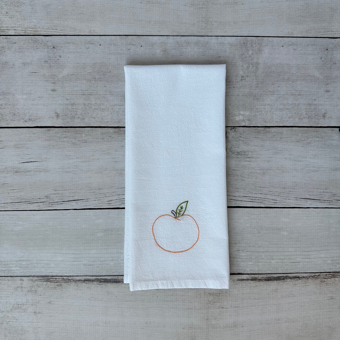 White tea towel with embroidered peach design on a wooden surface