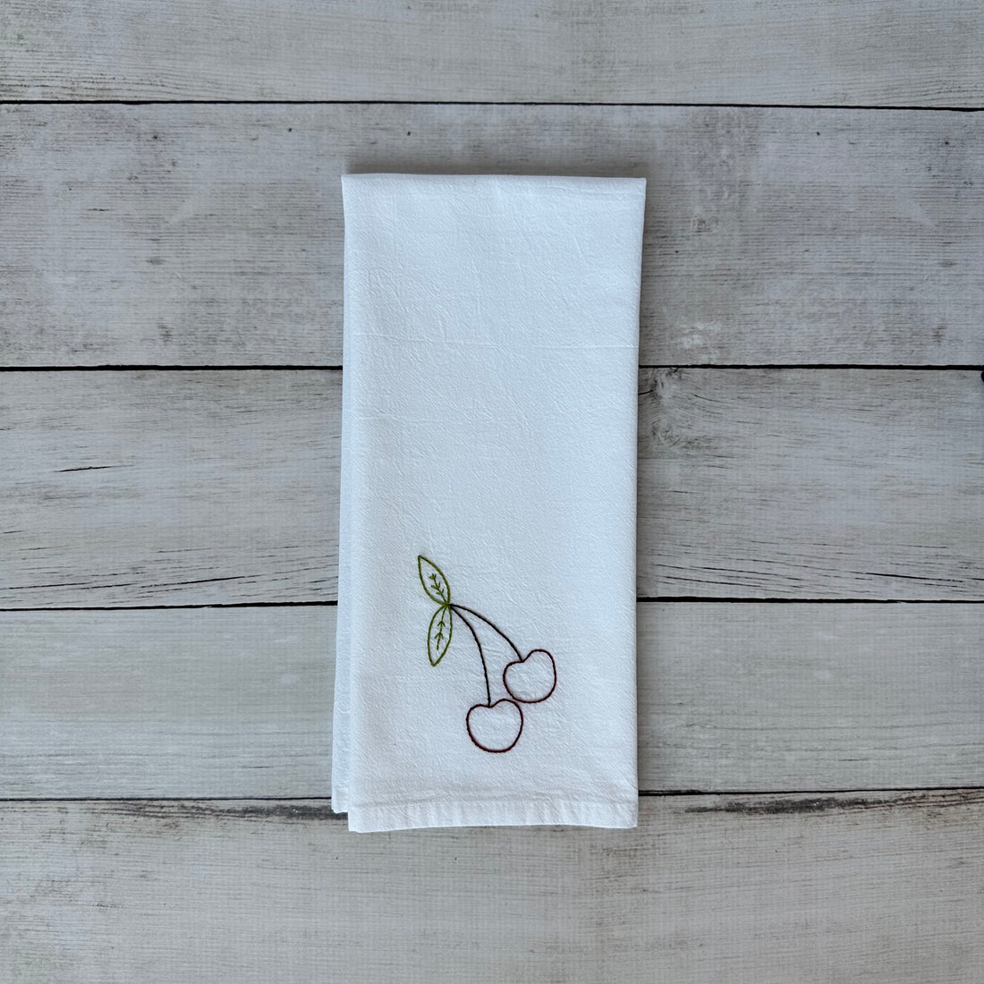 White tea towel with embroidered cherry design on a wooden surface