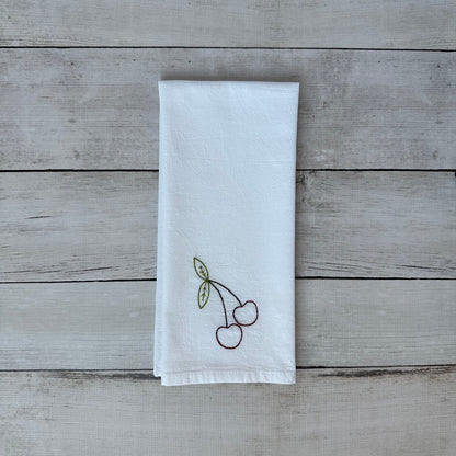 White tea towel with embroidered cherry design on a wooden surface